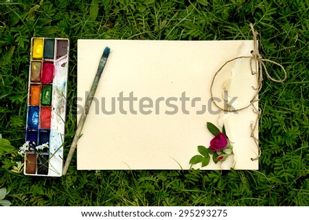 handmade drawing pad with flowers and leaves of wild rose, watercolor paints and brush on the background of grass