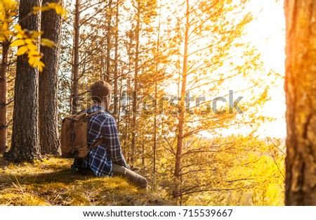 A young guy with a backpack sits on the edge of the cliffs and looks off into the distance at the forest, looking the autumn landscape, the sun's rays.