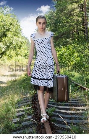 Girl in polka-dot dress with a suitcase in hand goes on the railroad tracks at the camera