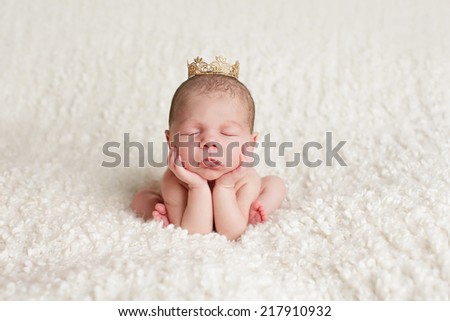 A newborn baby sits on a white blanket held up by its hands and elbows while wearing a gold crown