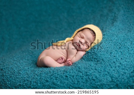 Newborn baby curled up on a blue blanket wearing a yellow knit cap
