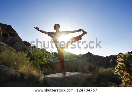 Man in the yoga pose Utthita Hasta Padangustasana (Extended Hand-To-Big-Toe Pose) while balanced in a desert landscape with the sun bursting through his core.
