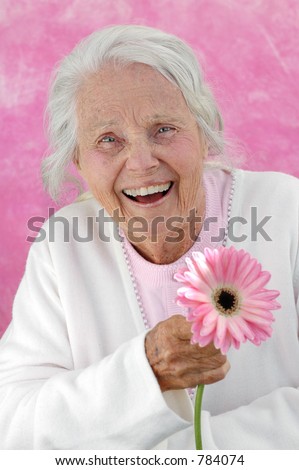 Laughing great grandmother with a pink Gerbera flower. Photographed on a pink background. Woman in her eighties.