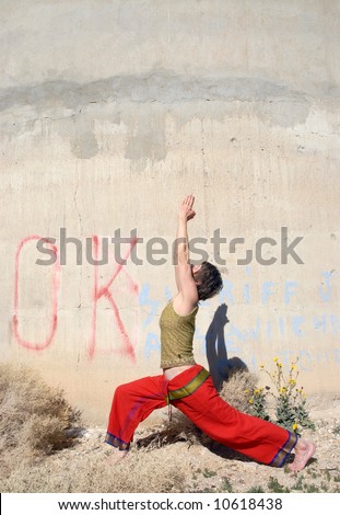 Woman in the yoga pose warrior 1, outdoors near a large industrial wall in the desert.