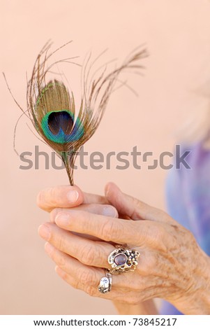 The adorned hand of a woman in her 80\'s holding a peacock feather.