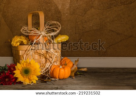 Autumn Basket A tabletop a basket of mini pumpkins and gourds, dried leaves, a sunflower, and red mums.