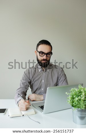 Man with beard in glasses taking notes with laptop and notepad looking on screen