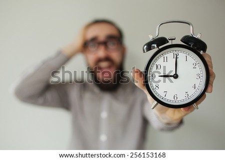 Man in panic with beard and glasses holds alarm clock and head scared of deadline