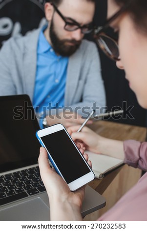 Young woman with smartphone taking notes over laptop in cafe with man in glasses