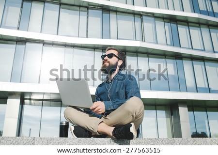 young hipster businessman with beard in sunglasses work with laptop outdoors in front of office building