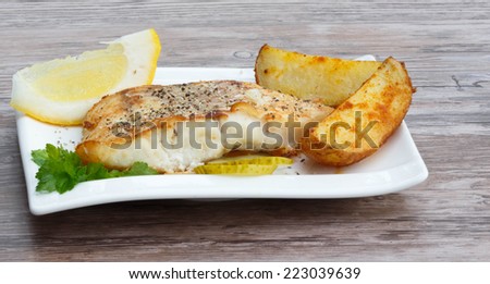 White fish with potato wedges, lemon and celery on white plat, wooden background
