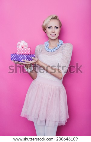 Beautiful blonde woman with cream colored dress holding pink and purple gift boxes. Studio portrait over bright pink background