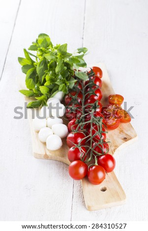 cherry tomatoes on branch and the tomatoes cut on slices, cheese mozzarella, fresh basil on wooden board and white table