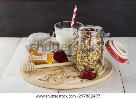 granola with nuts in glass jar, strawberry, glass of milk and jar of honey on white wooden table