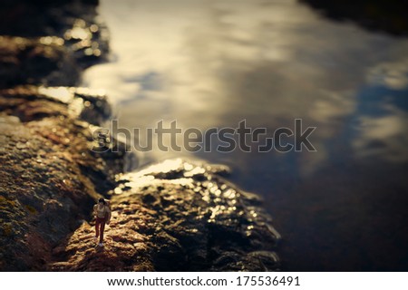 Female hiker on a rocky coast, sky reflecting in water behind her, miniature figure