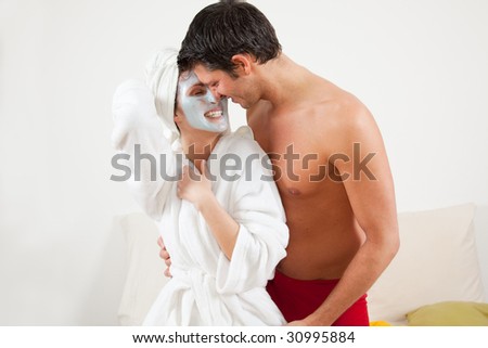 Kissing joyful couple with bathrobe and beauty mask making wellness at home
