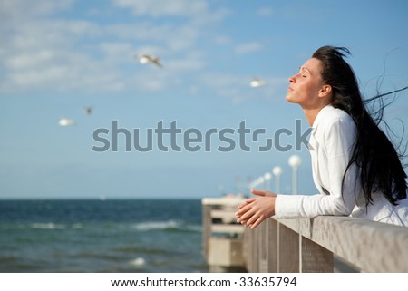 Brunette woman with flying winday hair on boardwalk looking the wide coast space with flying seagulls in background feeling free