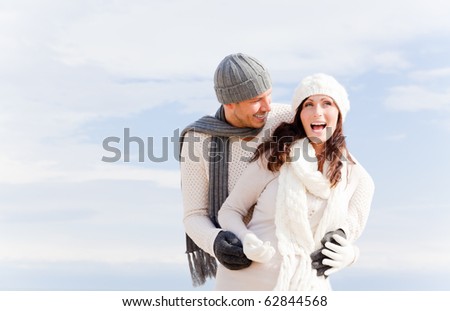 happy pair of male and female embracing and having fun wearing warm clothes outside on coast behind blue sky