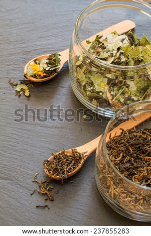Dry black and herb tea in wood spoons and glass jars on dark stone background