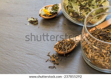 Dry black and herb tea in wood spoons and glass jars on dark stone background