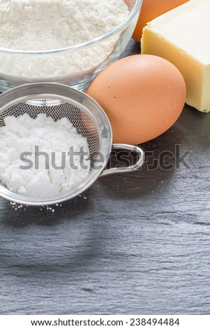 Baking ingredient - flour, eggs, butter and strainer with sugar powder on dark stone background