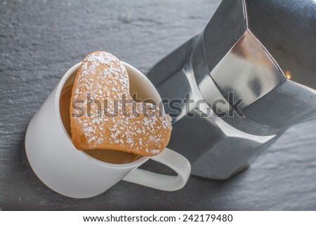 Cup of coffee with heart shaped cookie on top, coffee maker on dark stone background
