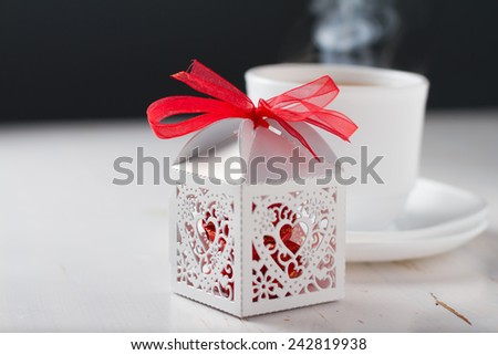 Present, white lacy box with red chocolate candies, cup od tea steaming on white wood background