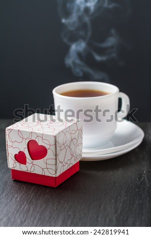 Present, chocolate candies box with hearts, cup od tea steaming on dark stone background