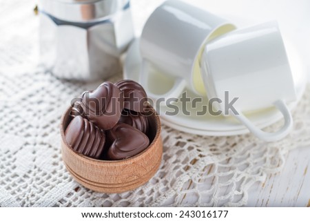 Chocolate candies in wood bowl, coffee cups, coffee maker on white knitted tablecloth, wood background