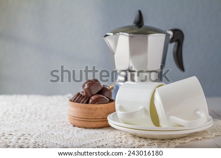 Chocolate candies in wood bowl, coffee cups, coffee maker on white knitted tablecloth, wood background