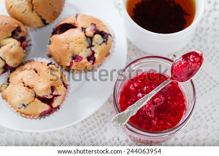 Raspberry jam in a spoon, berry muffins and black tea on white knitted tablecloth, top view