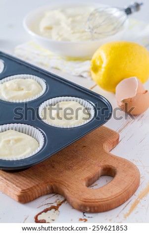 Lemon cupcakes preparation - cupcakes in forms, egg shells, dough in white bowl, wire whisk, white wood background