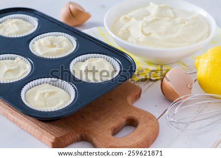Lemon cupcakes preparation - cupcakes in forms, egg shells, dough in white bowl, wire whisk, white wood background