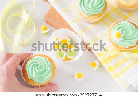 Spring cupcakes preparation - hand holding cupcake, sugar flowers, ribbon, plaid napkin, white wood background, top view