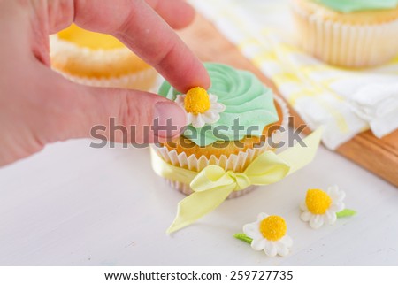 Spring cupcakes preparation - hand holding cupcake, sugar flowers, ribbon, plaid napkin, white wood background