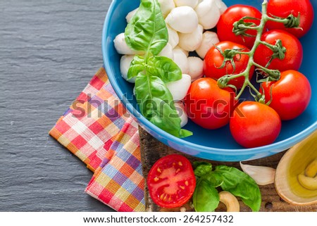 Caprese salad ingredients - cherry tomatoes, mozzarella, basil leafs in blue bowl, olive oil, nuts, chili, garlic, plaid napkin, dark stone background, top view, closeup
