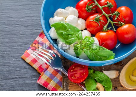Caprese salad ingredients - cherry tomatoes, mozzarella, basil leafs in blue bowl, olive oil, nuts, chili, garlic, plaid napkin, dark stone background, top view, closeup
