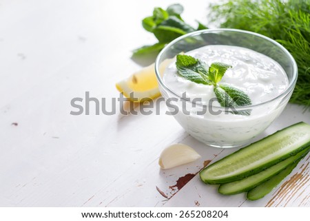Tzatziki sauce in glass bowl, with ingredients - cut cucumber, mint, dill, lemon, garlic, white wood background