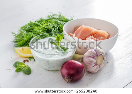 Tzatziki sauce in glass bowl, with ingredients - cut cucumber, mint, dill, lemon, garlic and meat for souvlaki, white wood background