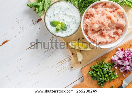 Tzatziki sauce in glass bowl, with ingredients - cut cucumber, mint, dill, lemon, garlic and meat for souvlaki, white wood background, top view