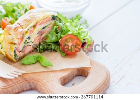 Strudel pie with salmon and spinach, served on wood board with salad, cherry tomatoes, sour cream, white wood background, closeup