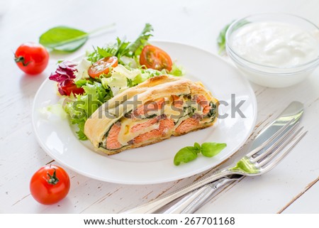 Strudel pie with salmon and spinach, served on white plate with salad, cherry tomatoes, sour cream, white wood background