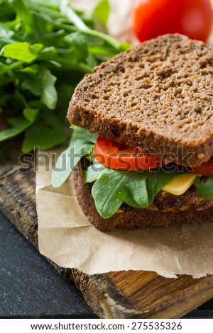 Vegetarian sandwich with zucchini burger, cheese, ruccola and tomatoes, wood board, dark stone background, closeup