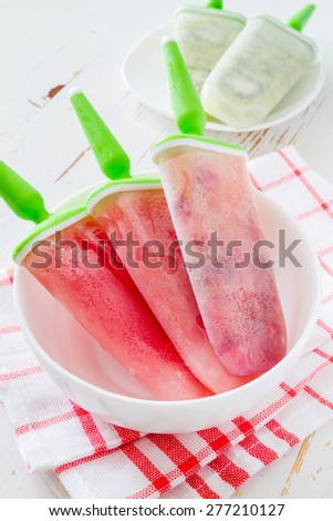 Fruit ice pops (watermelon, berry, orange) in white bowl, plaid napkin, white wood background