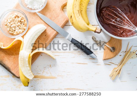 Banana pops preparation - banana, chocolate, nuts, coconut powder, white wood background, top view