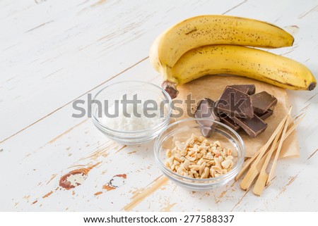 Banana pops preparation - banana, chocolate, nuts, coconut powder, white wood background