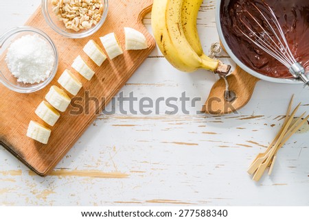 Banana pops preparation - banana, chocolate, nuts, coconut powder, white wood background, top view
