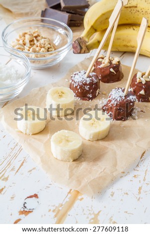 Banana pops preparation - banana, nuts, coconut powder, chocolate, baking paper,white wood background