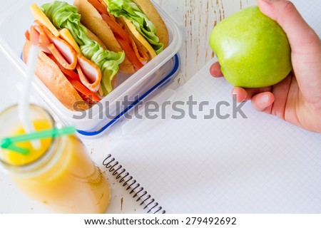 Eating lunch on the workplace - notepad, pen, sandwich, hand holding apple, orange juice, white wood background, top view
