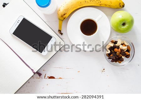 Breakfast - notepad, pen, coffee, yogurt, nuts, banana, apple, phone, white wood background, top view